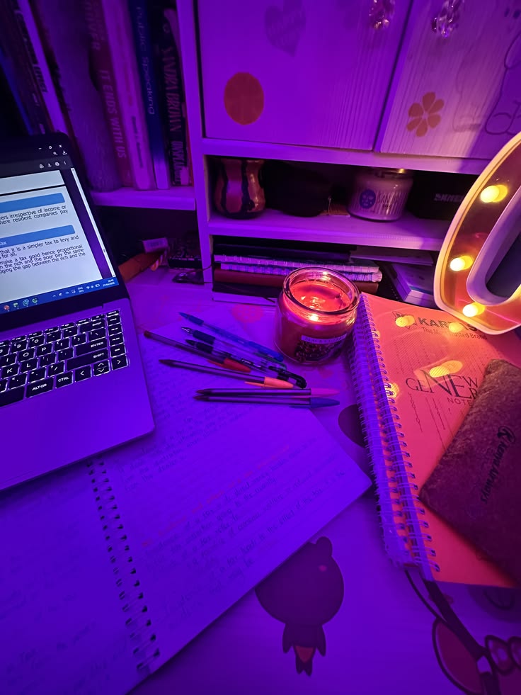 A desk at night in purple light: a laptop, open notebooks, pens and a glowing candle